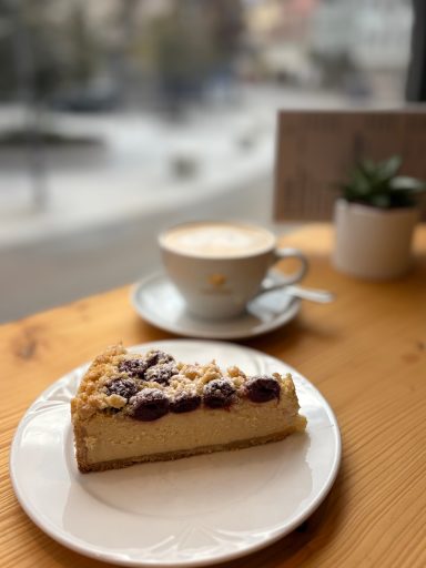 Foto von einem Käsekuchen mit Kirschen und Streuseln. Eie Tasse gefüllt mit leckerem Milchkaffee.