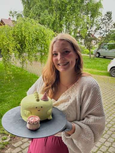 kleine grüne Torte, dekoriert als Dinosauerier mit Partyhütchen und Minitorte mit einer 19 darauf. Gehalten wird sie vom Geburtstagskind.