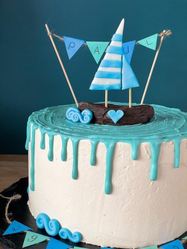 Eine weiße Torte mit modelliertem Boot. Mit blauen Akzenten aus Schokolade, Wellen und einem Wimpel mit dem Namen des Taufkindes